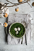 Top view of a beautifully laid Easter table with a bowl filled with grass and speckled eggs, surrounded by old silver cutlery and a branch with hanging eggs. A soft, textured background complements the festive spring theme.