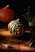 An artistic still life captures the rustic autumn atmosphere with textured pumpkins, bright herbs and a marigold on a wooden table. The warm lighting emphasises the cosy, seasonal ambience.