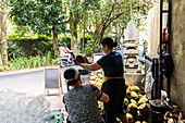 Two women open coconuts on a busy tropical street. The scene is surrounded by greenery and motorbikes and captures the essence of daily life and local culinary traditions.