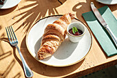 A golden croissant rests on a plate next to a small bowl of jam on a sunlit wooden table A fork and knife lie next to it, emphasising the cosy breakfast scene