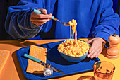 A cosy scene with a person enjoying a bowl of creamy macaroni and cheese. The rich, melted cheese lies on the fork, while the table is coloured in bright blue and yellow.