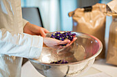 A person in a lab coat holds dried mallow flowers over a metal bowl and demonstrates the preparation process for a natural infusion. Brown paper bags can be seen in the background.