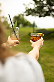 Two hands raise glowing cocktails in the open air, capturing a moment of relaxation One hand holds a drink garnished with leaves, the other a citrus-embellished one