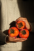 A woman gently cradles three ripe persimmons against a textured, cosy jumper background. The warm colours contrast beautifully with the fabric and exude an autumnal mood.