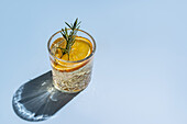 Ein gekühlter Zitruscocktail, garniert mit einem Rosmarinzweig und einer Orangenscheibe, serviert in einem alten Kristallglas vor einem hellblauen Hintergrund, der einen sanften Schatten wirft.