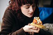 A young adult woman with curly red hair enjoys a delicious slice of pizza and concentrates on her food in a relaxed environment.