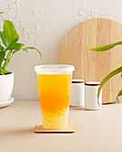 A large cup of iced tea in bright orange stands on a coaster, surrounded by green plants and simple white decor that creates a cosy and modern aesthetic.