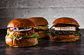 Drei Gourmet-Burger mit saftigen Patties, geschmolzenem Käse, Zwiebeln, knackigem Salat, Speck und weichen Brötchen. Präsentiert auf einer dunklen Fläche vor einem rustikalen Hintergrund.