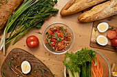 A lively table with crusty baguette, fresh vegetables, hard-boiled eggs and a salad in a glass bowl, showing the preparation of a healthy meal.