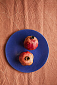 Two ripe pomegranates lie on a bright blue plate on a textured brown fabric background. The contrast highlights the natural colour of the fruit and gives them a visual appeal.