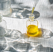 Eine Glasflasche mit goldenem, natürlichem Körperöl steht in sanftem Sonnenlicht, während eine Pipette weitere Flüssigkeit hinzufügt. Es wird aus Pflanzen wie Calendula, Rose, Lavendel und Arnika hergestellt und dient der Hautpflege und dem Wohlbefinden.