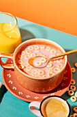 A lively breakfast scene with a bowl of muesli with pink milk, accompanied by a glass of orange juice and a cup of coffee, against a colourful background.