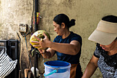 Two women skilfully open a coconut and let the water run into a blue bucket. The scene is set against a rustic wall and emphasises traditional techniques and teamwork.