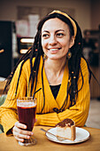 A cheerful woman with dreadlocks and a yellow jumper enjoys a slice of cake and a drink in a cosy café, radiating happiness and relaxation.