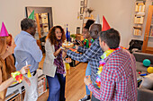A lively birthday party with friends gathered around a cake in a cosy room. Festive decorations, colourful hats and cheerful smiles create a joyful atmosphere.