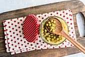 A wooden spoon scoops homemade organic peas from an open jar. The bright green contrasts with a red dotted lid and a cloth, all on a rustic wooden board that creates a cosy kitchen atmosphere.
