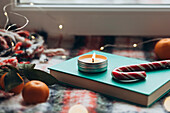 Cosy Christmas arrangement with a lit candle, a candy cane and mandarins on a book, surrounded by fairy lights and a warm blanket, embodying the spirit of Christmas warmth and joy.