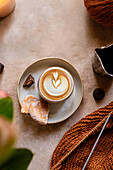 A warm scene with a knitted fabric next to a creamy latte art coffee, accompanied by biscuits and chocolate, on a rustic surface for a cosy autumn atmosphere.