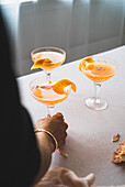 Cut-out, unrecognisable person holding a glass with a bright tangerine cocktail, garnished with an orange peel, on a laid table. This image represents a perfect blend of luxury and refreshment.