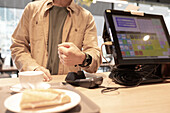A customer in a café uses a contactless payment method at the counter with a smartwatch A cup of coffee and a muffin can be seen in the foreground, with a digital screen and a card reader in focus