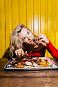 A woman enjoys a delicious barbecue platter with different types of meat against a bright yellow background. The scene captures the joy of eating and the lively atmosphere.