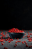 A dark bowl with bright red peppercorns stands against a textured, dark grey background. Scattered peppercorns surround the bowl, creating a contrast that emphasises its rich, pink colour.