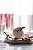 A stylish Easter decoration with a decorative egg with feathers on a pastel-coloured tablecloth, complemented by elegant pink glasses.