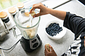 Ein Kind gibt Bananenscheiben in einen Mixer für einen frischen Smoothie, daneben eine Schale mit Blaubeeren - perfekt für ein nahrhaftes Frühstück oder einen Snack, der Gesundheit und Geschmack vereint