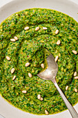 Top view of a rich, bright green pesto sauce with pine nuts and red pepper flakes, served in a white bowl with a silver spoon that captures the essence of authentic Italian cuisine.
