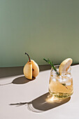 A refreshing summer cocktail, decorated with a slice of pear and a sprig of rosemary, against a colourful green background with artistic shadows.