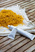 Two white tubes of marigold flower cream lie on a wooden base next to a pile of dried yellow marigold flowers. A simple, natural and holistic beauty concept.