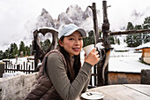 An Asian woman enjoys a hot drink on the Geisleralm in the Italian Dolomites Surrounded by snow-capped peaks and rustic wooden decorations, she savours the tranquil, picturesque surroundings