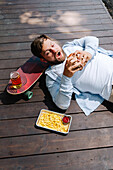 A man enjoys a burger, fries and a drink while relaxing on an outdoor wooden terrace. A skateboard nearby gives the scene a relaxed, adventurous atmosphere.