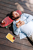 A man is lying on a wooden terrace enjoying a burger with a tray of fries and a drink on a skateboard next to him. The scene captures a relaxed outdoor barbecue atmosphere.