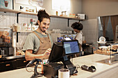 A cheerful café employee in an apron smiles as she operates the till at the counter. Shelves of cups and pastries can be seen in the cosy café.