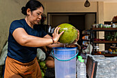 A woman expertly opens a fresh coconut and pours the water into a container. The scene captures the lively atmosphere of a local market and emphasises traditional skills.
