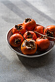 Bowl of fresh, ripe persimmons, freshly picked in the orchard, presented on a subtly textured grey surface, with natural sunlight highlighting their rich orange colour.