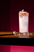 A stunning Paloma cocktail with a salted rim sits on a wooden surface surrounded by ice cubes. The bright pink background emphasises the refreshing look of this classic tequila drink