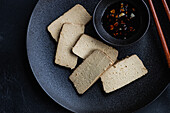 Top view of savoury tofu and soy cheese slices arranged on a dark plate, accompanied by spicy pepper sauce in a small bowl and a side dish of rice, served with chopsticks on a textured black background.