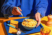 A person in a blue jumper enjoys a bowl of macaroni and cheese on a bright blue tray. A yellow dotted napkin complements the colourful surroundings and conveys warmth and cosiness.