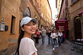 A smiling Asian tourist in a white T-shirt and cap walks through the charming narrow streets of Volterra, Tuscany Blurred background with locals and tourists enjoying the historic architecture