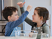 Two young siblings wearing star-patterned aprons play happily together in a kitchen. A mixing bowl can be seen in the foreground, indicating a cooking activity.