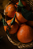 Eine Nahaufnahme, die saftige Orangen mit leuchtend grünen Blättern in einem rustikalen geflochtenen Korb zeigt. Die Orangen, die vor Frische glänzen, verkörpern Vitalität und natürliche Schönheit.