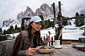 A quiet moment on the Geisleralm, Dolomites, Italy, with an Asian woman enjoying a meal with snow-capped peaks in the background Cosy meal in a breathtaking mountain landscape