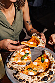One person holds a slice of fresh Margherita pizza, topped with mozzarella and basil. The pizza is accompanied by a chilled lemonade. A casual outdoor dining experience.