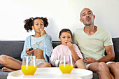 A family sits on a sofa and watches a film together. Two children and an adult are enjoying popcorn and there are glasses of orange juice on the table, creating a cosy living room atmosphere.
