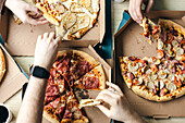 Top view of a group of unrecognisable friends gathered around a table sharing various slices of pizza from cardboard boxes. The image captures a casual and friendly dining situation.