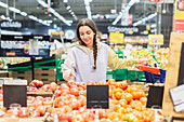A woman in a casual hoodie buys fresh tomatoes in a busy supermarket and carefully selects the best ones to prepare her meal.