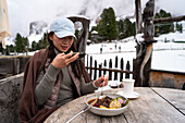 An Asian person enjoys a hearty meal at the Geisleralm, surrounded by snow-capped peaks in the Dolomites, Italy The cosy atmosphere offers a wonderful alpine experience