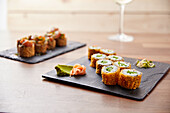 An elegant arrangement of sushi rolls on a black slate, with crispy rolls and hosomaki, complemented by wasabi and pickled ginger. A blurred glass of white wine can be seen in the background.